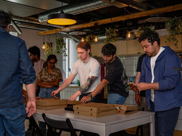 A group of coworkers gather around a table in an office common area or kitchen to grab slices of pizza from cardboard boxes.