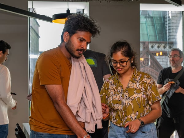 A candid photograph of a group of people indoors, featuring a man in a brown t-shirt and a woman in a patterned shirt looking down, while others stand in the background.