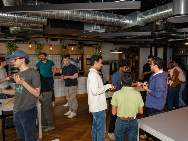 A group of young professionals socializing and eating pizza in a modern office breakroom with exposed ceiling pipes.
