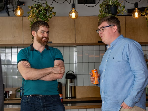 Two men are standing and chatting in an office kitchen or breakroom, with one holding a can of Fanta and the other standing with arms crossed.