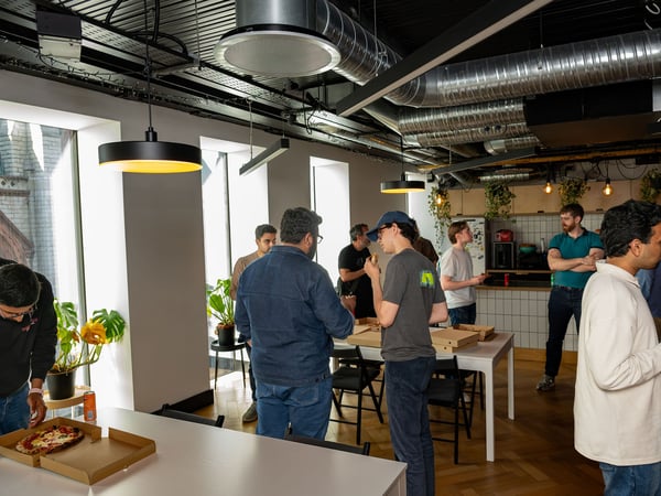 A group of colleagues gathering in a modern office breakroom to socialize and eat pizza.