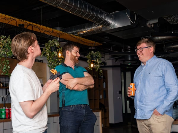 Three men are standing and socializing in an office breakroom or kitchen area, sharing a laugh while holding drinks and a snack.