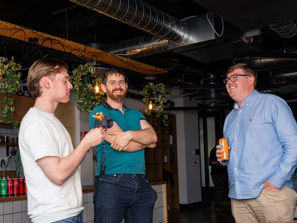 Three men are standing and socializing in an office breakroom or kitchen area, with one holding a can of Fanta and another holding a small snack.