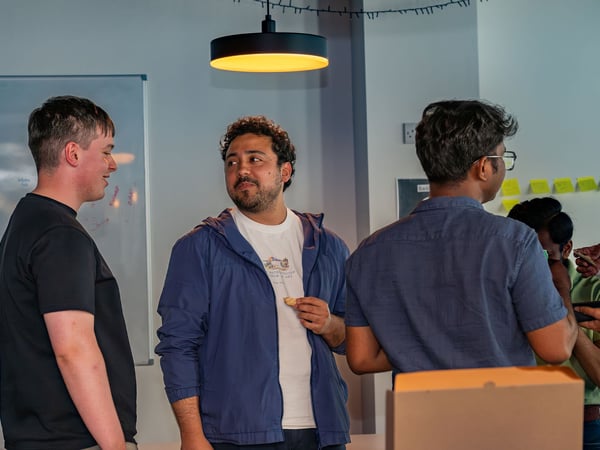 A group of young men are standing and conversing in an office or workshop setting, with a whiteboard and a warm hanging light in the background.