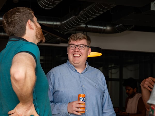 A candid photograph of a group of men socializing indoors, with one man in a light blue shirt and glasses laughing while holding a Fanta can.
