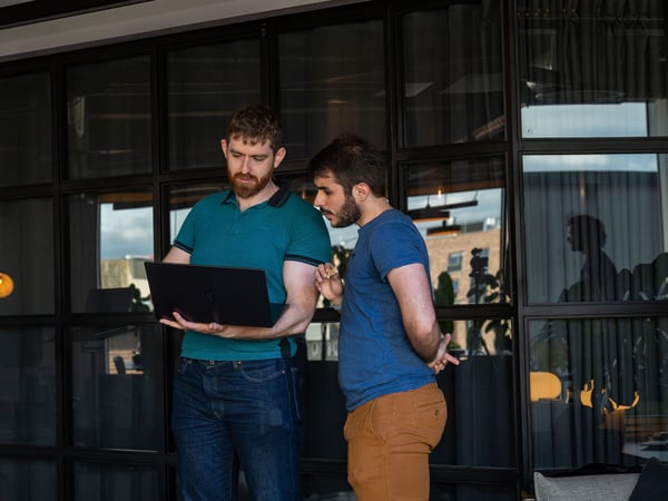 Two men standing in front of a large gridded glass window, focused on a laptop screen that one of them is holding.