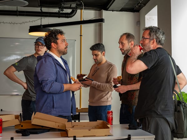 A group of colleagues standing together in an office setting, chatting while eating pizza and drinking sodas during a casual break.