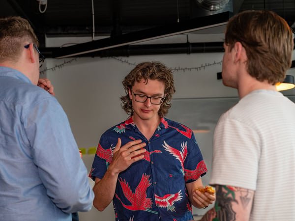 A candid photograph of three young men engaged in a casual conversation indoors, with the central figure wearing a blue shirt patterned with red cranes.