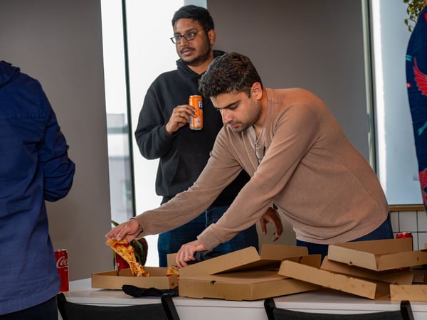 A candid indoor photograph showing a group of colleagues gathering around a table with open pizza boxes, with one man reaching for a slice of pizza while another holds a can of Fanta.