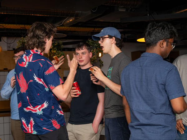 A group of young men are socializing, eating pizza, and drinking sodas in an indoor breakroom or office kitchen setting.