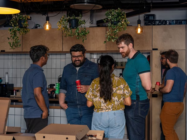 A group of colleagues socializing and chatting in an office kitchen or break room during an event with pizza and drinks.