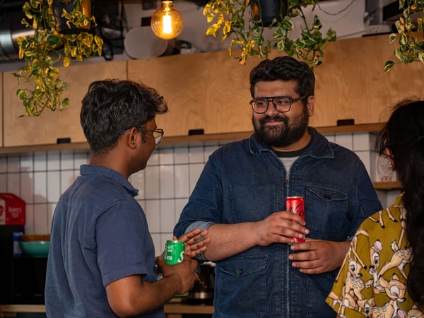A candid photo of people socializing in an indoor kitchen or cafe setting with hanging plants and warm lighting, focusing on two men conversing while holding soda cans.