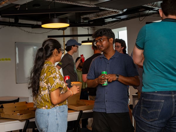 A group of people are seen socializing and chatting indoors at an office event, with pizza boxes on a table and some individuals holding drinks.