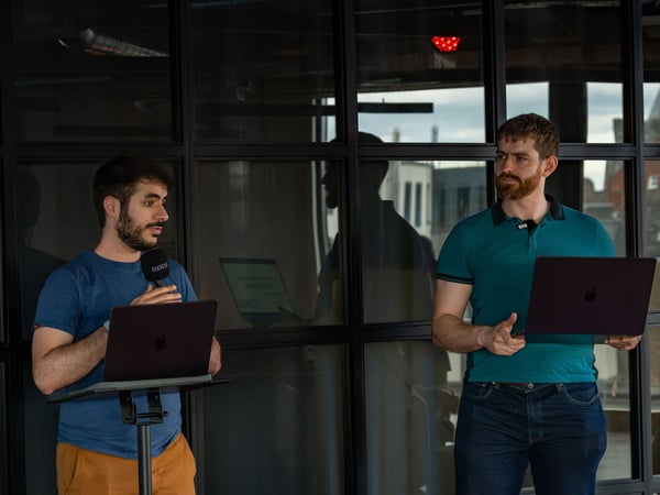 Two men are standing indoors in front of a dark glass partition, both holding laptops. The man on the left is speaking into a microphone on a stand, while the man on the right holds his laptop and looks in his direction.