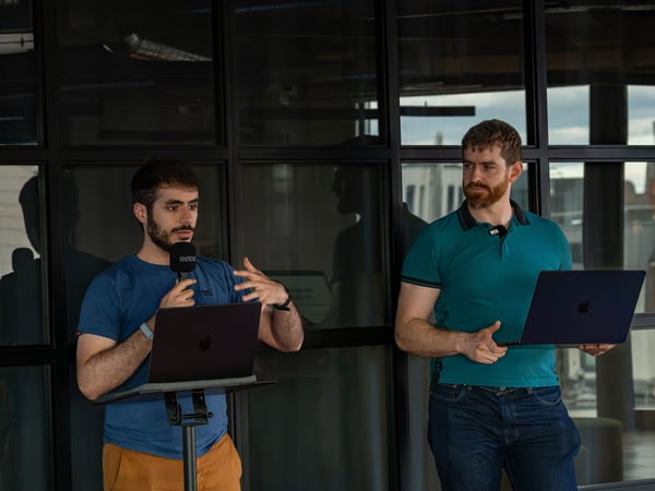 Two men are presenting indoors in front of a dark glass paneled wall; the man on the left is speaking into a RØDE microphone while gesturing, and the man on the right holds a laptop and listens.
