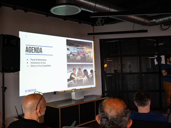 A photograph showing an indoor presentation event where an audience is watching a projected slide titled 'AGENDA' with details about an AI Tinkerers meetup.