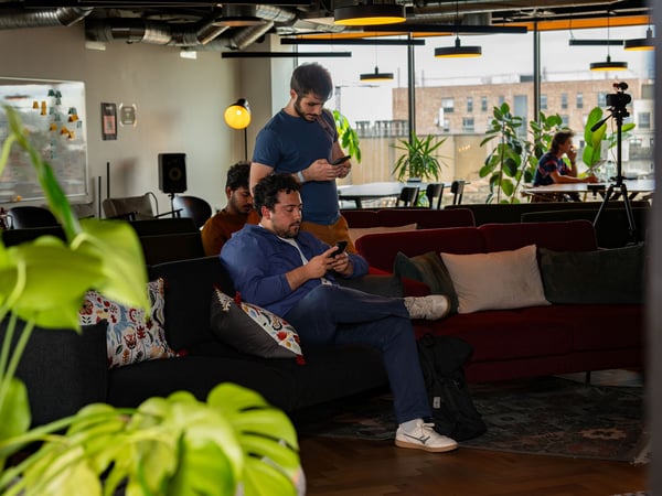 A photograph of a modern co-working space where several men are sitting on sofas and looking at their smartphones, surrounded by indoor plants and office decor.