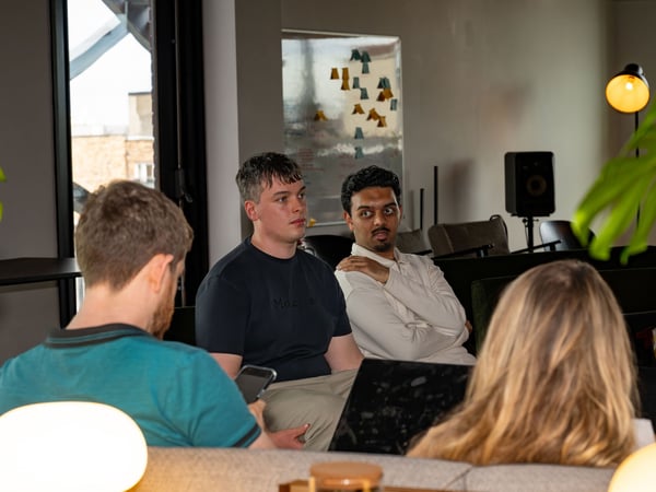 A group of people are gathered in a modern indoor lounge or office setting, with two men in the middle ground looking attentively towards the left, while two others in the foreground are seen from behind.