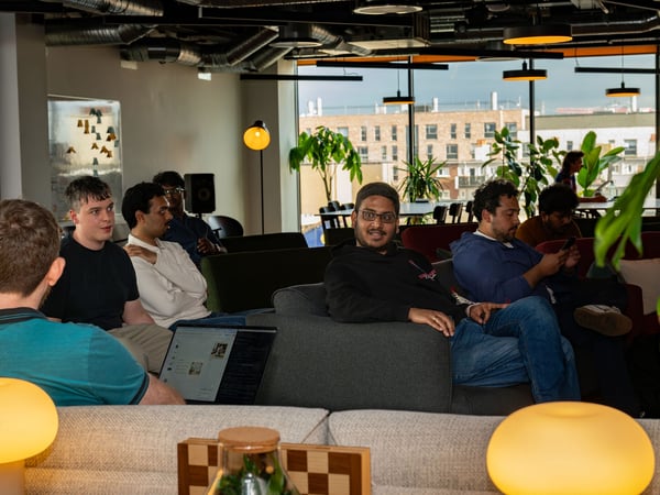 A group of people are gathered in a modern office lounge, sitting on sofas and engaged in a casual meeting or discussion. Warm mushroom-shaped lamps glow in the foreground while large windows in the background show adjacent buildings.