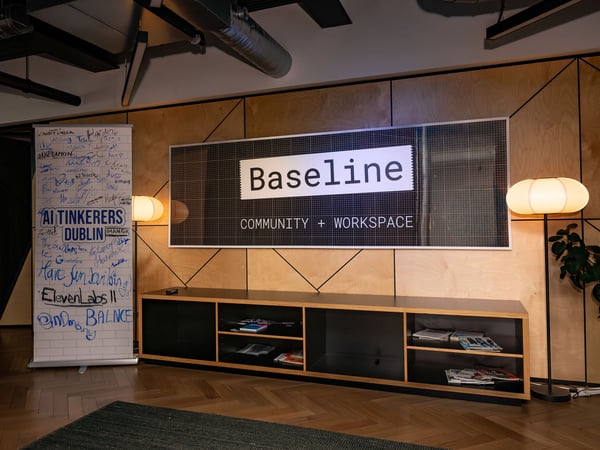 An indoor photograph of a modern workspace interior featuring a large 'Baseline COMMUNITY + WORKSPACE' sign on a wooden paneled wall, flanked by warm floor lamps and a banner filled with signatures.