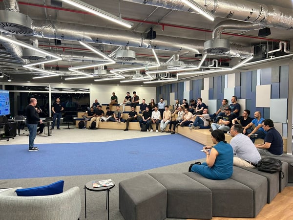 A photograph capturing a presentation or internal meeting in a modern, open-plan office space, featuring a speaker addressing a large audience seated on tiered wooden benches and modular gray furniture.