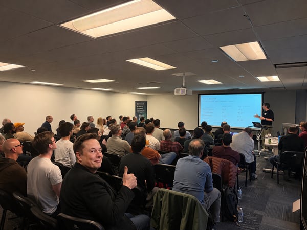 A photograph of a presentation inside a conference room, featuring Elon Musk in the foreground turning around to give a thumbs-up to the camera while an audience watches a slideshow presentation.