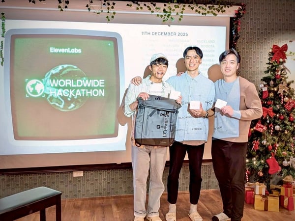 Three young men stand together, posing for a photo in an indoor setting, likely during an awards ceremony for the ElevenLabs WORLDWIDE HACKATHON, which is displayed on a large projection screen behind them, next to a decorated Christmas tree.