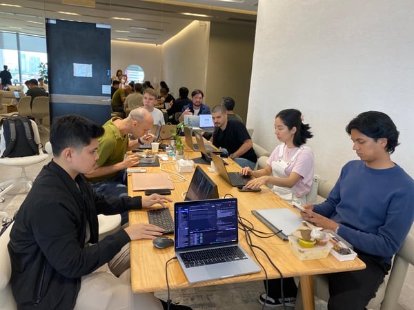 A group of people are working on laptops and eating at a large wooden table in a modern, brightly lit coworking space.