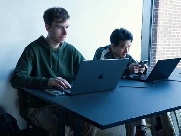 Two young men are sitting at a dark desk in a modern indoor space; one is typing on a laptop while the other looks at a smartphone next to a second laptop.