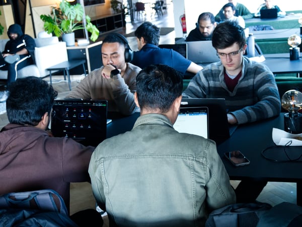 A group of young men are focused on their laptops while working in a modern, indoor shared workspace or cafe.