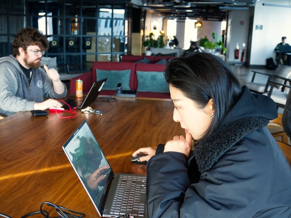Two people are focused on their laptops while sitting at a large wooden table in a modern, open-plan co-working space.