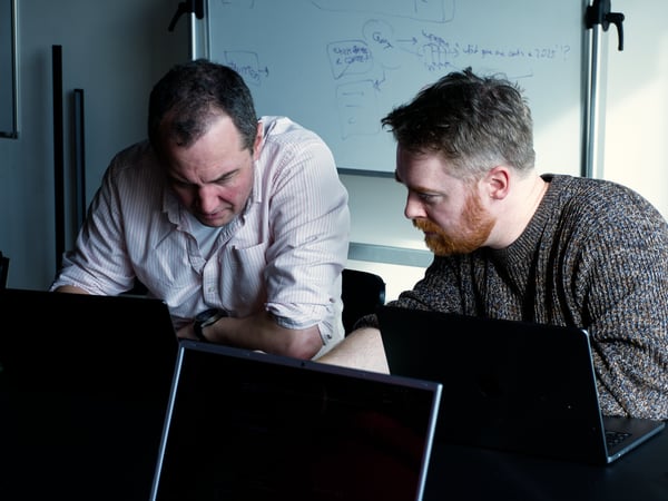 Two men are intently focused on a laptop screen in an office environment, with a whiteboard featuring diagrams and notes in the background.