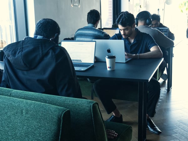 A group of individuals are focused on their laptops in a modern coworking space or cafe, with one man facing the camera while others have their backs turned.
