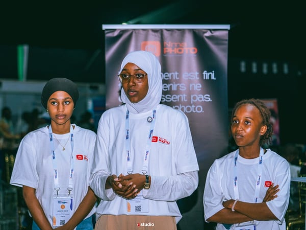 Three young women wearing matching white t-shirts and lanyards stand in front of a promotional banner at an event.