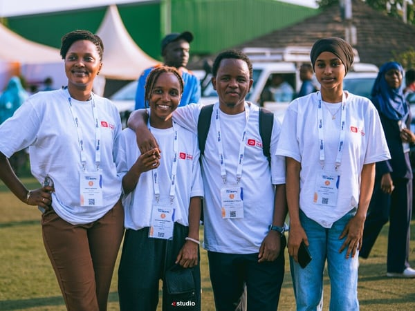 A group of four young people pose together outdoors at an event, wearing matching white t-shirts and event lanyards.