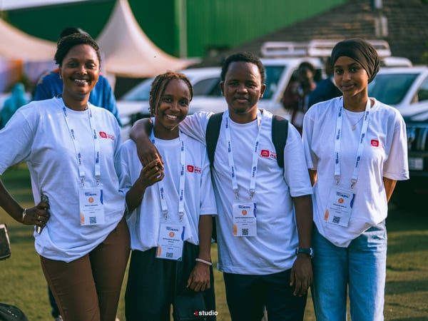 A group of four young people, three women and one man, posing together outdoors at an event. They are all wearing white t-shirts with matching lanyards and identification badges.