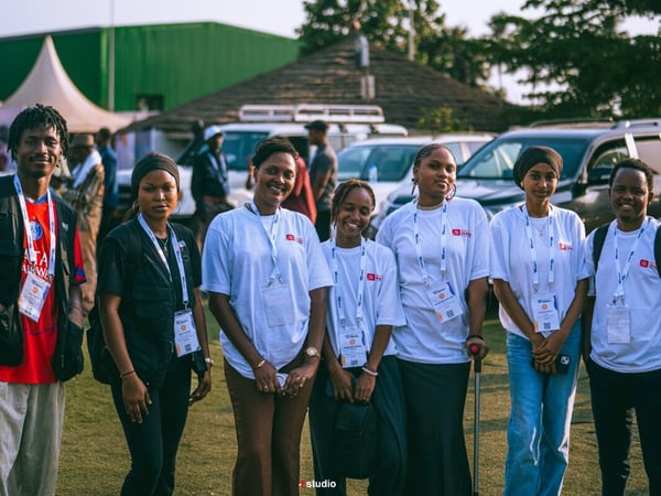 A group of seven young people posing for a photo outdoors at an event, wearing lanyards and casual clothing.