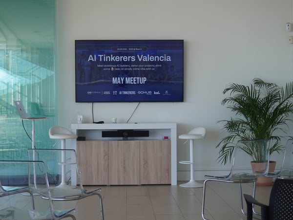 An indoor shot of an empty presentation room set up for an 'AI Tinkerers Valencia' meetup, featuring a large screen and transparent chairs.