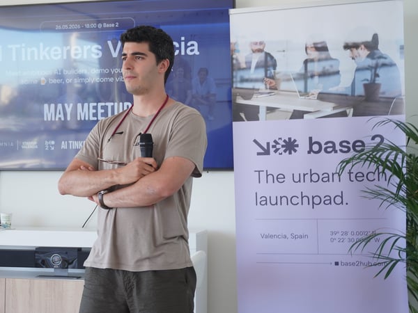 A man stands with arms crossed holding a microphone at an indoor tech meetup, in front of a presentation screen and a banner.
