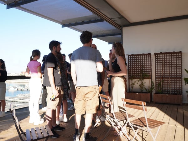A group of people socializing on a sunny outdoor terrace overlooking a harbor.