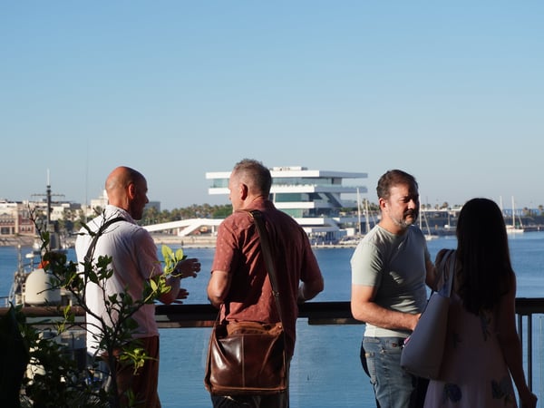 A group of people standing on a terrace or balcony, socializing while overlooking a harbor with a modern building in the background.