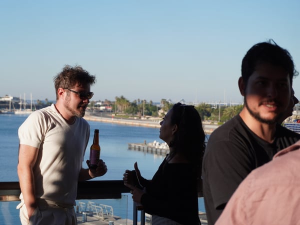 A group of people socializing outdoors on a terrace overlooking a scenic marina under a clear blue sky.