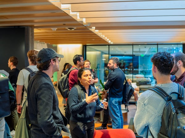 A group of people are gathered indoors in a modern office lobby or event space, engaged in conversation. A woman in a dark blue sweater is gesturing and speaking to a man wearing a baseball cap in the foreground.