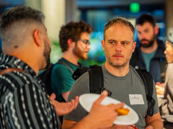 A candid photograph of people socializing at an indoor event, featuring a man named Lukas wearing a nametag and listening to another person gesturing with a plate of food.