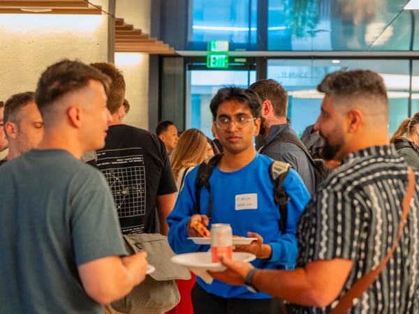 A group of people are seen socializing and networking at an indoor event, with some individuals holding plates of pizza and drinks.