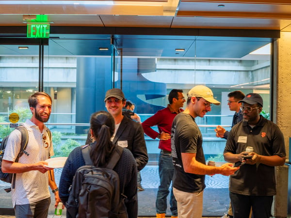 A group of people are seen socializing and conversing indoors near a glass entrance with an illuminated green exit sign.