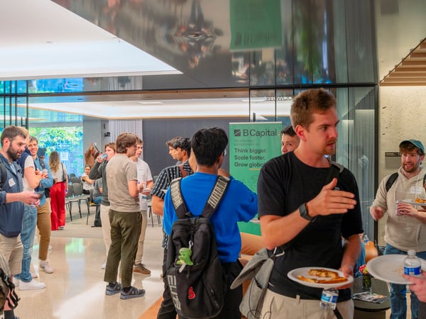 A group of people are socializing, eating, and networking in a modern, well-lit indoor lobby or event space featuring a B Capital banner.