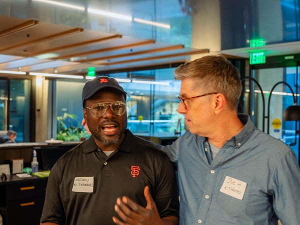 A photograph of two men standing indoors at an event, wearing name tags that read 'ABDOU AI Tinkerers' and 'Joe H AI Tinkerers'.