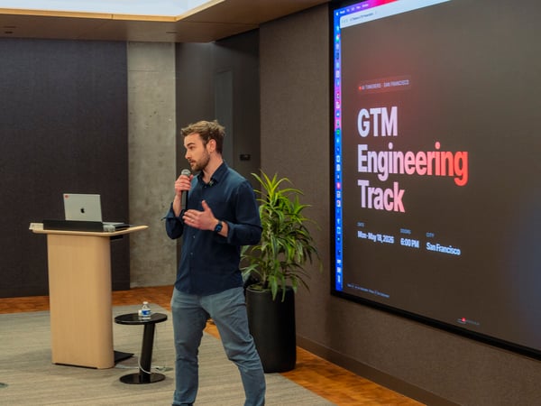 A man stands on a stage speaking into a microphone next to a large projection screen displaying event details for 'GTM Engineering Track' by AI Tinkerers in San Francisco.