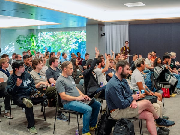 A photograph showing an audience of people seated in a modern conference room, with several individuals raising their hands during a presentation or Q&A session.